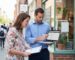 A professional local SEO consultant performing an on-site audit of a Google Business Profile for a small urban storefront, using a laptop and checklist to discuss optimizations with the business owner.