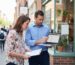 A professional local SEO consultant performing an on-site audit of a Google Business Profile for a small urban storefront, using a laptop and checklist to discuss optimizations with the business owner.