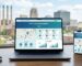 Modern digital workspace with laptop displaying website design interface and analytics dashboard, Akron skyline visible through window, smartphone mirroring site
