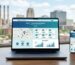Modern digital workspace with laptop displaying website design interface and analytics dashboard, Akron skyline visible through window, smartphone mirroring site