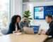 Three marketing consultants collaborating around a conference table in a bright modern office, reviewing an AI-driven SEO dashboard on a laptop while a wall monitor displays website mockups and SEO graphs